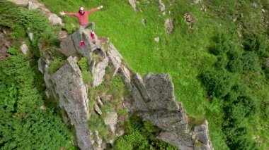 Yürüyüşçünün hava manzarası büyük bir kayanın üzerinde, aşağıdaki sisli, pürüzlü uçurumların ve yeşil vadinin nefes kesici manzarasının tadını çıkarıyor. Kırmızı kapüşonlu ve bandanalı, dingin, rüzgarlı manzaraya renk katıyor..