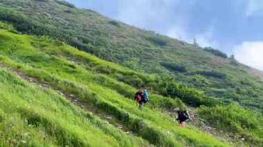 İki yürüyüşçü parçalı bulutlu gökyüzünün altında yeşil dağ yolu boyunca yürürler. Yuvarlanan tepeler ve uzak tepeler nefes kesici bir zemin oluşturuyor. Yürüyüşçüler iyi donanımlı, temiz ve manzaralı yolculuğun tadını çıkarıyorlar..
