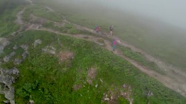 Üç yürüyüşçünün hava manzarası sisli dağ sırtı boyunca yüksek kaya oluşumlarıyla çevrili kuvvetli rüzgarla yürüyor. Kayalık arazi ve yemyeşil bitki örtüsü dingin bir atmosfer yaratır.