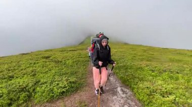 Üç yürüyüşçü sisli dağ yolunda yürür. Her biri büyük bir sırt çantası taşıyor ve destek için yürüyüş direkleri kullanıyor. Bereketli yeşillikler arasında rüzgarlar eser, sis dingin ve mistik bir ortam yaratır..