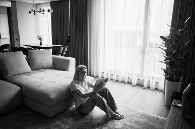 Kanepenin yanında oturan ve kitap okuyan genç bir kadın. Güzel bir kadın, ahşap zeminde dinleniyor. Kitapla oturan kadın pencerenin karşısında, doğal ışıkta. Siyah ve beyaz resim.