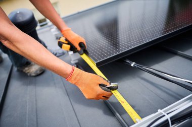 Fotovoltaik etkilerle elektrik üretmek için evin çatısına güneş paneli inşa eden işçiler. Montaj ekipmanlarını ölçmek için cetvel kullanan adamı kapat.