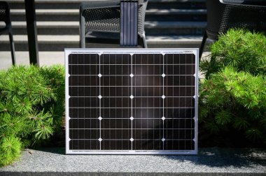 Fotovoltaik güneş paneli taştan çıkıntının üzerindeki canlı yeşil çalılara karşı modern, kentsel manzarada güneş ışığını emiyor. Yenilenebilir enerjinin şehir ortamlarına entegrasyonu kavramı.