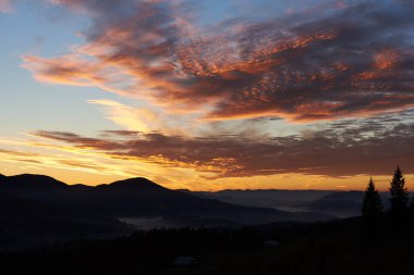 Nefes kesen günbatımı gökyüzünü pembe, turuncu, mor ve mavi gökyüzüne harmanlanmış canlı renklerle boyar. Siluetli dağlar, çam ağaçları dingin bir zemin oluşturur. Huzurlu ve resmedilmiş manzara.