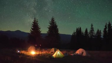 Dağlarda gece kampı. Ormanlı dağ manzarasının ortasında iki çadır parlak, yıldızlarla dolu gökyüzünün altında kamp ateşinin yanında sıcacık parlıyor. Samanyolu 'nun altındaki kamp alanında dinlenen turistlerin siluetleri.