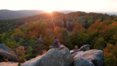 Ukrayna 'daki Karpat Dağları' ndaki Dovbush Kayalıkları 'ndaki kaya oluşumunun üzerinde oturan dağcı görüntüsü. Güneş batıyor, sonbahar ormanları ve uzak tepeler üzerinde parıldıyor, nefes kesici bir manzara yaratıyor..