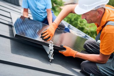 İşçiler evin çatısına fotovoltaik güneş paneli inşa ediyorlar. Kasklı ve eldivenli erkek teknisyenlerin güneş modülünü açık alandaki hex anahtarı yardımıyla kurmalarını sağlayın. Yenilenebilir enerji.