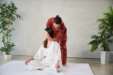 Masör Tayland yogası masajı yapıyor. Kadın terapist, dirsek kullanarak kadın hastaya geleneksel masaj yapıyor. Müşteri mindere oturuyor, meditasyon yapıyor, sakin bir atmosferde el masajının keyfini çıkarıyor..