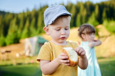 Sarı tişörtlü ve mavi şapkalı genç, çimenli kamp alanında ayakta dururken ellerindeki çöreği dikkatle inceliyor. Açık mavi elbiseli kız arka planda duruyor, etrafında çam ağaçları ve çam ağaçları var..