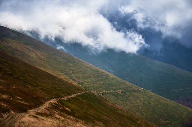Dik dağ yamaçlarında yemyeşil çimenler ve kahverengi bitki örtüsüyle kaplı yamaçtan yukarı doğru uzanan dolambaçlı toprak yollar var. Yukarıda, kalın bulutlar kısmen belirsiz görünüyor, gizem ve dram hissi ekliyor..