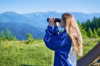 Çimenli çayırlarda duran kadın, uzak dağları gözlemlemek için dürbün kullanıyor, mavi kapüşonlu bir şey giyiyor. Sahne, yemyeşil tepelerin ve açık mavi gökyüzünün zeminine kurulur..