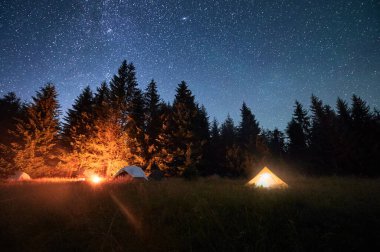 Çimenli, berrak, yıldızlı gökyüzünün altında parlayan kamp ateşiyle aydınlatılmış birkaç çadır. Dağlarda yüksek çam ağaçlarıyla çevrili. Doğada kamp yaparak geçirilen gecenin huzurlu güzelliği..
