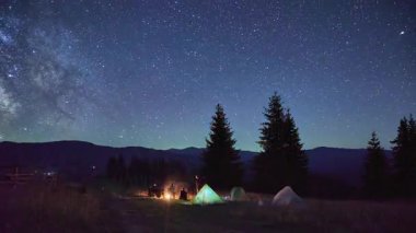 Dağlarda gece kampı. Ormanlı dağ manzarasının ortasında iki çadır parlak, yıldızlarla dolu gökyüzünün altında kamp ateşinin yanında sıcacık parlıyor. Samanyolu 'nun altındaki kamp alanında dinlenen turistlerin siluetleri.