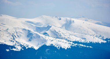 Karla kaplı dağ zirvesi açık mavi gökyüzünün altında, yamaçlarda kar parçaları ve dağınık ağaçlarla. Sakin kış manzarası aşağıdaki karanlık ve ormanlık alanlarla keskin bir tezat oluşturuyor. Chornohora, Ukrayna