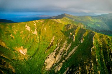 Dramatik gökyüzünün altındaki engebeli dağ zirvelerinin nefes kesen yeşil manzarası. Manzara, canlı manzarayla karamsar bulutlar arasındaki kontrastı yakalar, güneş ışığının kırılışıyla. Karpatlar, Kornogora