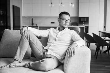 Evindeki kanepede dinlenen yakışıklı bir adam. Yakışıklı genç erkek düşünceli bir şekilde başka tarafa bakıyor. Modern mutfağın arka planında gündelik giysiler giymiş bir adam yatıyor. Siyah ve beyaz resim.