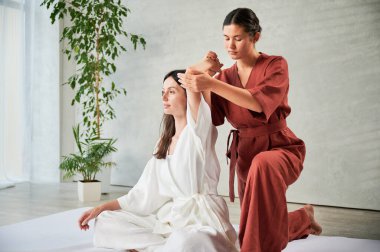 Masör Tayland yogası masajı yapıyor. Kadın terapist, kadın hasta için evde geleneksel masaj tedavisi uyguluyor. Müşteri mindere oturuyor, meditasyon yapıyor, sakin bir atmosferde el masajının keyfini çıkarıyor..