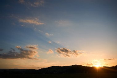 Güneş ufukta batar ve altın parıltısını yeryüzüne saçar. Yumuşak bulutlar, sıcak renklerle aydınlatılmış, açık mavi gökyüzüne karşı yüzerler. Huzurlu bir sahne, günün son ışığı yavaşça kayboluyor..