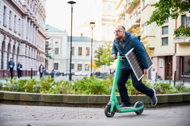 Adam güneş paneli taşırken elektrikli scooter kullanıyor. Elektrikli scooterlar için sürdürülebilir enerji kaynağı olarak güneş enerjisinin entegrasyonu, çevre dostu kentsel ulaşımı teşvik etmek.