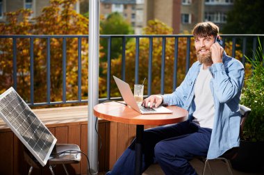Adam dizüstü bilgisayarla çalışıyor ve dışarıdaki masada bir bardak buzlu kahveyle akıllı telefon kullanıyor. Yakınlardaki güneş paneli, teknoloji ve sürdürülebilirliği birleştiren sürdürülebilir, modern çevre dostu çalışma alanını vurguluyor.