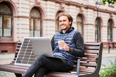 Şehirdeki ahşap bankta oturan adam elinde bir fincan kahveyle dizüstü bilgisayarla çalışıyor. Güneş paneli, tarihi mimarinin arkaplanının ortasında sürdürülebilir, hareketli çalışma alanının vurgulandığı,.