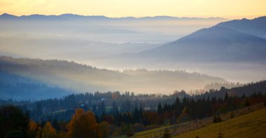 Sabah güneşi, ormanlık tepelerin arasına kurulmuş sakin dağ köyünü aydınlatan sislerin arasından akıyor. Işık, sonbahar yapraklarına ve yamaçlara saçılmış evlere nazikçe dokunan ruhani ışınlar yaratır..