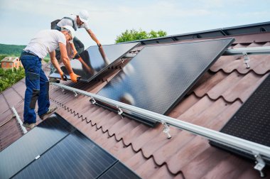 İşçiler evin çatısına güneş paneli inşa ediyorlar. Dışarıda fotovoltaik güneş modülü taşıyan kasklı iki adam var. Alternatif, yeşil ve yenilenebilir enerji üretimi kavramı.