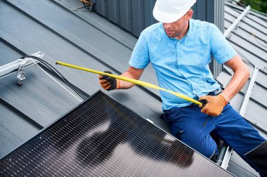 Fotovoltaik etkilerle elektrik üretmek için evin çatısına güneş paneli inşa eden işçiler. Kurulum için montaj ekipmanlarını ölçmek için cetvel kullanan adam.