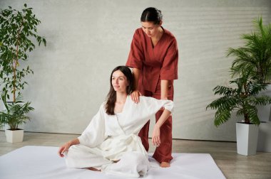 Masör Tayland yogası masajı yapıyor. Kadın terapist, kadın hasta için evde geleneksel masaj tedavisi uyguluyor. Müşteri mindere oturuyor, meditasyon yapıyor, sakin bir atmosferde el masajının keyfini çıkarıyor..