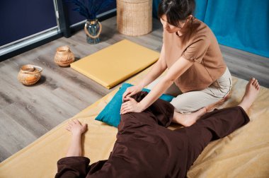 Masör Tayland yogası masajı yapıyor. Geleneksel masaj tedavisi uygulayan kadın terapist, kadın hastaların vücudunu geriyor. Müşteri paspasın üzerinde uzanıyor, sakin atmosferde el masajının keyfini çıkarıyor..