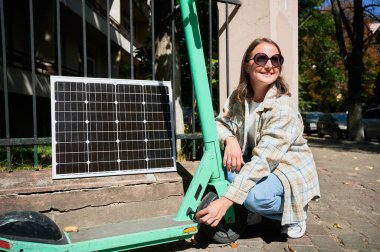 Güneş panelini elektrikli scooter 'a bağlayan kadın sürdürülebilir yenilenebilir enerji uygulamasının altını çiziyor. Çevre dostu teknolojinin günlük taşımacılığa entegrasyonu kavramı.