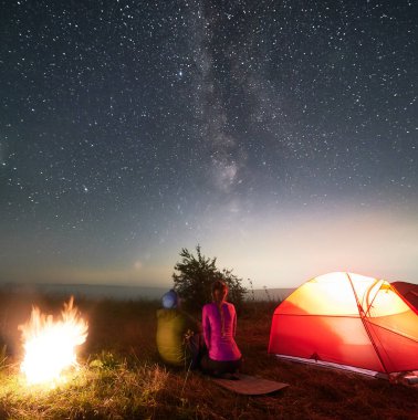 Şenlik ateşinin yanında oturan ve yıldızlı güzel gökyüzüne bakan kadın ve erkek yolcuların arka manzarası. Gece yıldızlı gökyüzü çimenli çayırların üzerinde şenlik ateşiyle aydınlatılmış kamp çadırı ve yürüyüşçülerle.