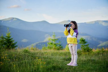 Genç kız çimenli çayırda duruyor, dürbünle bakıyor, sarı ve mor kazak giyiyor. Arkasında, yuvarlanan yeşil tepeler ve dağlar açık mavi gökyüzünün altında uzanıyor..