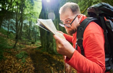 Sakallı turist yakın plan haritası incelerken sık ormandaki büyük kayaların önünde duruyor. Gri sırt çantası ve gözlüklü gezgin, açık hava macerası, yürüyüş ya da tırmanma rotası planlıyor..