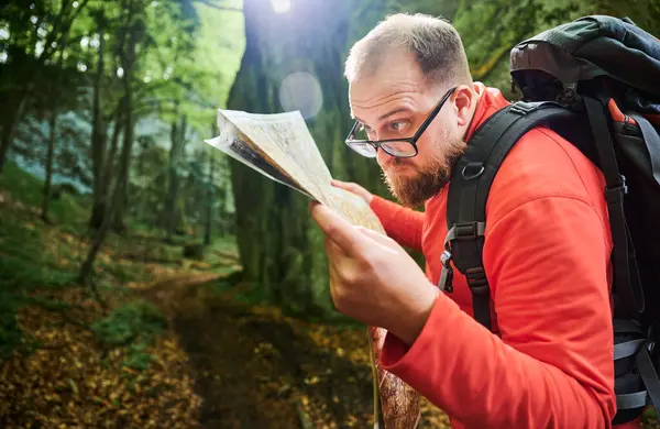 Sakallı turist yakın plan haritası incelerken sık ormandaki büyük kayaların önünde duruyor. Gri sırt çantası ve gözlüklü gezgin, açık hava macerası, yürüyüş ya da tırmanma rotası planlıyor..