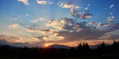 Gün batımında mavi ve yumuşak pembe bulutların renkleriyle huzurlu bir akşam göğü. Ufuk hafifçe parlıyor, siluetli çam ağaçları ve yuvarlanan tepeler üzerine ışık saçıyor, huzurlu ve geniş bir manzara yaratıyor..