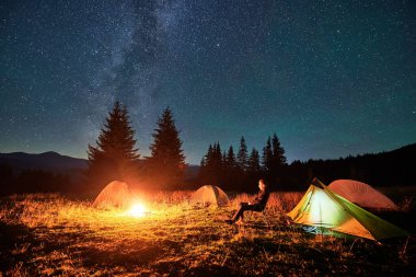 Yürüyüşçü, muhteşem yıldızlı gökyüzünün altında parlayan çadırlarla çevrili kamp ateşinde dinleniyor. Samanyolu uzanıyor, huzurlu dağ yabanda huzurlu ve büyülü bir atmosfer yaratıyor..