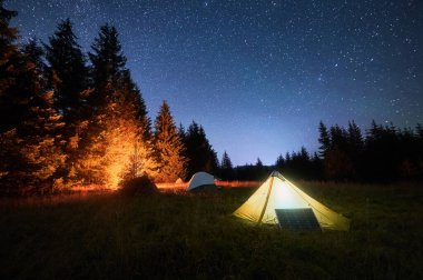 Yellow tent beside solar panel under stunning, star-filled night sky. Nearby, other tents and trees are lit by warm light from campfire, highlighting eco-friendly and serene camping experience.