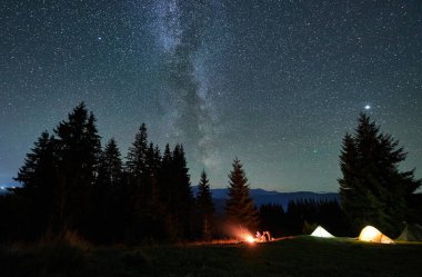 Dağlarda gece kampı. Ormanlı dağ manzarasının ortasında iki çadır parlak, yıldızlarla dolu gökyüzünün altında kamp ateşinin yanında sıcacık parlıyor. Samanyolu 'nun altındaki kamp alanında dinlenen turistlerin siluetleri.