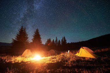 Samanyolu ile yıldızlı gökyüzünün altında çimenli açıklıklarda parlayan kamp ateşi etrafında birkaç çadır. Kamp ateşinin sıcacık ışığı olay yerini aydınlatıyor, dağlarda konforlu ve huzurlu bir gece kamp ortamı yaratıyor..