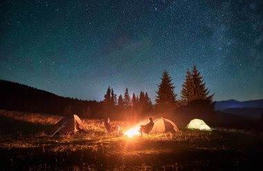Kamp alanındaki aydınlık çadırların yakınında yıldızlarla kaplı gökyüzünün altında kamp ateşinde dinlenen çift yürüyüşçülerinin silueti. Dağlarda gece kampı, gece gökyüzü büyülü ve huzurlu bir atmosfer yaratır..