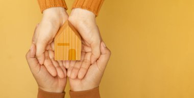 Sarı ahşap bir evi tutan, sevgiyi, evi simgeleyen ve sıcaklığı ve birliği ile bir aile kuran iki el.