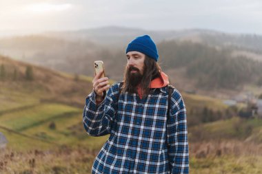 Akıllı telefondan haritayı kontrol eden gezgin, tepede mavi bere, ekose gömlek, turuncu kazak ve gün batımında omuzlarında deri bir sırt çantasıyla yürürken. Sırtında sırt çantası olan yürüyüşçü bir erkek tırmanıcı arka planda duruyor.