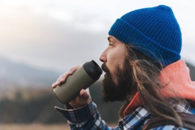 Erkek gezgin gün batımının tadını çıkarırken resimli tepelerde yürüyor. Yürüyüş sırasında molada içen yürüyüşçü erkek, güzel bir dağ manzarasının arka planında durup, uzaklığa bakar..  