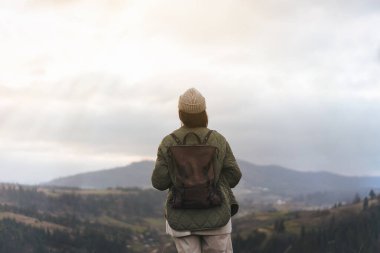 Sırt çantalı ve bereli bir kızın sırtından uçurumun kenarında durup manzaranın, gün batımının, dağların ve bulutların keyfini çıkarın..