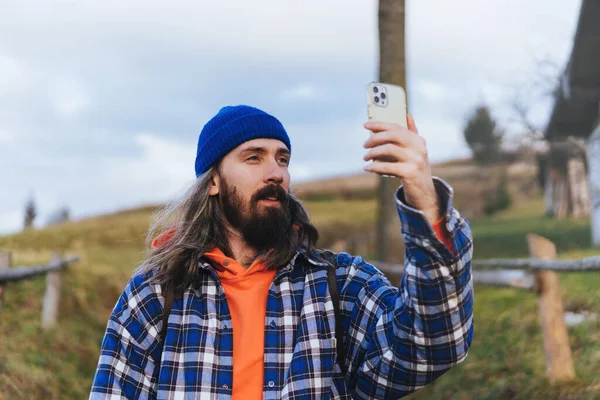 男ハイカーは山を歩いている間にスマートフォンで写真を撮る 青い帽子とジャケットを着て長い髪を持つ魅力的な男の肖像画 男の肖像画のライフスタイル — ストック写真
