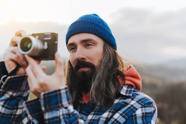 美しい丘を背景に青いビーニーで長い髪を持つ魅力的な旅行者の男の肖像画 古いカメラで日没の写真を撮る写真家旅行者 — ストック写真