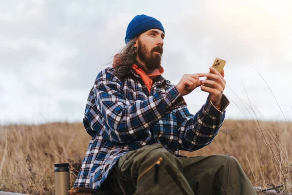 青い上着を着た若い髭の男の旅行写真家のフロントビュー日没の秋の田園地帯でトレッキング中にぼやけた緑の丘に対して座っている間にスマートフォンで写真を撮る — ストック写真