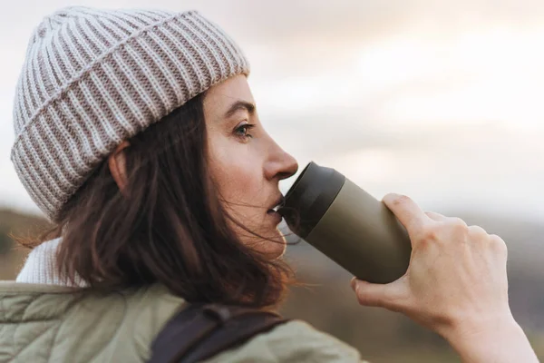 stock image Woman drinking water while standing on the hills and enjoying sunset during hiking in countryside. Female mountaineer looking at beautiful view with a water bottle in hand.