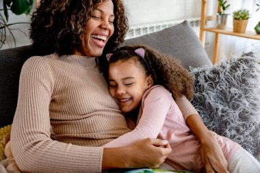 Afro-Amerikan anne ve kızı kanepede sarılıyorlar.